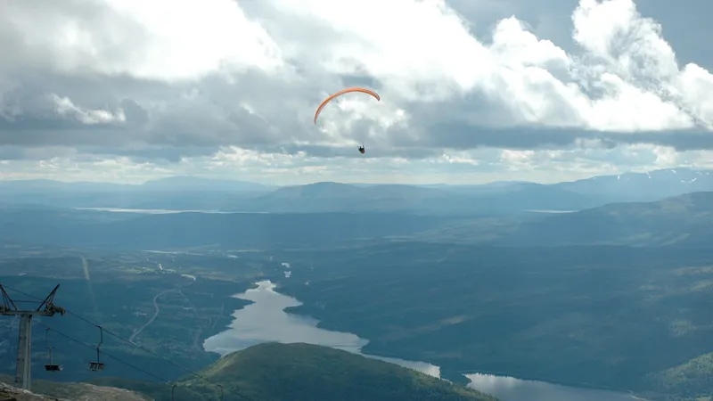 En person som flyger skärm högt upp i luften. Det är moln och fjäll i bakgrunden och i dalen en älv.