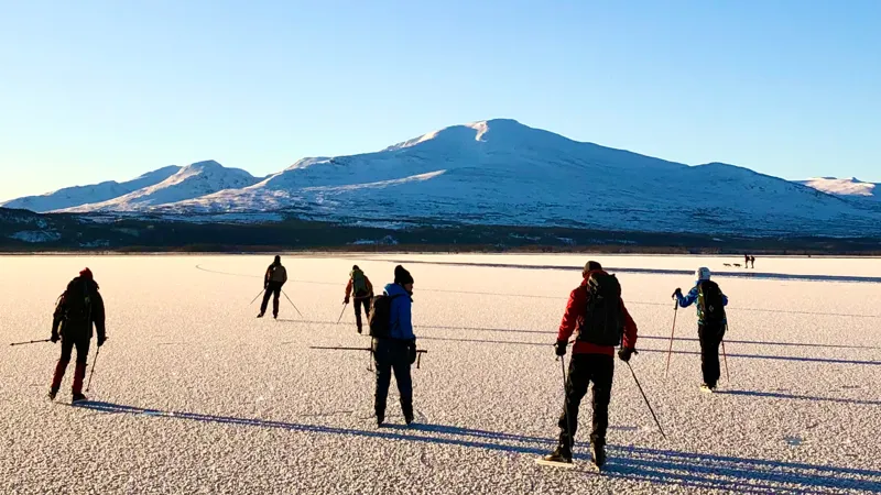 En grupp människor som åker skridskor på en sjö. Blå himmel och vita fjäll i bakgrunden.