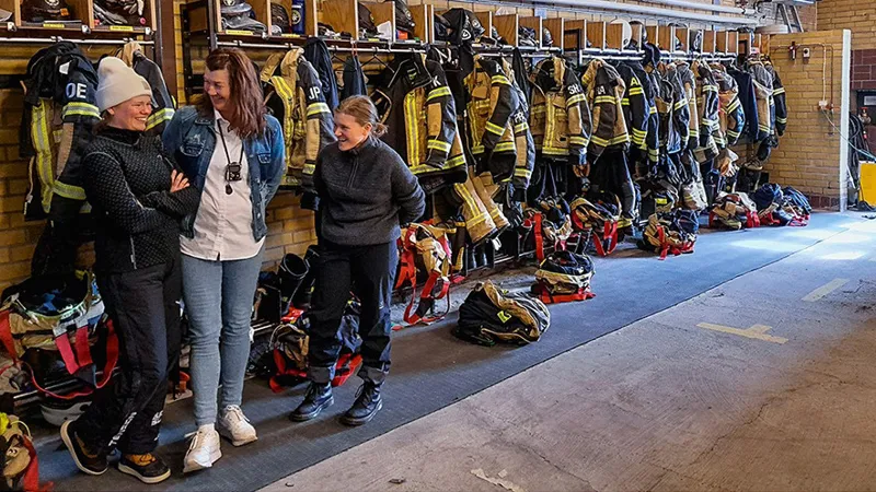 Tre personer är i en brandstation. Bakom dem hänger brandmännens kläder på rad.