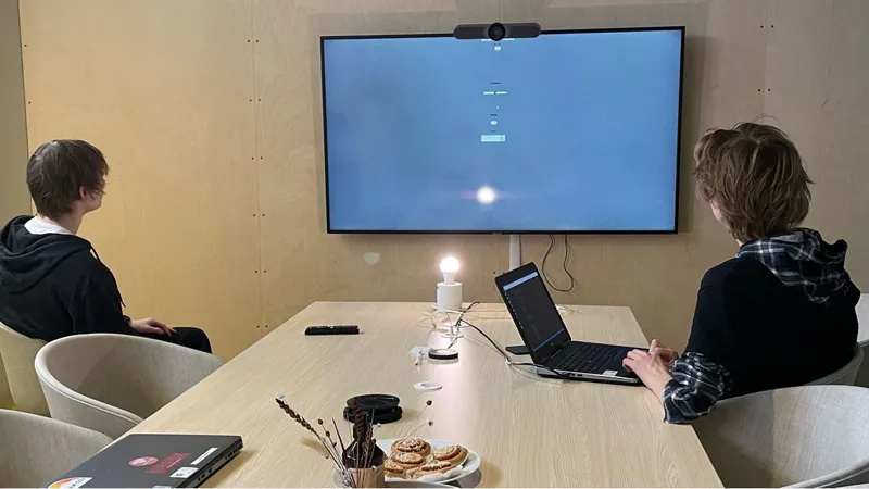 Två personer sitter vid ett bord med bärbara datorer och tittar på en stor skärm som sitter på väggen framför dem.