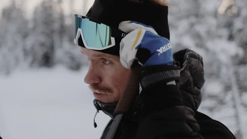 Närbild på ansiktet på en man som är ute på en skidtur. Oskarp snöig skog i bakgrunden.
